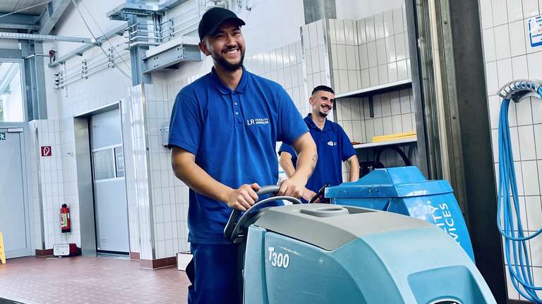 Ein Mitarbeiter in blauer Arbeitskleidung fährt lachend eine Bodenreinigungsmaschine, ein Kollege steht im Hintergrund daneben.