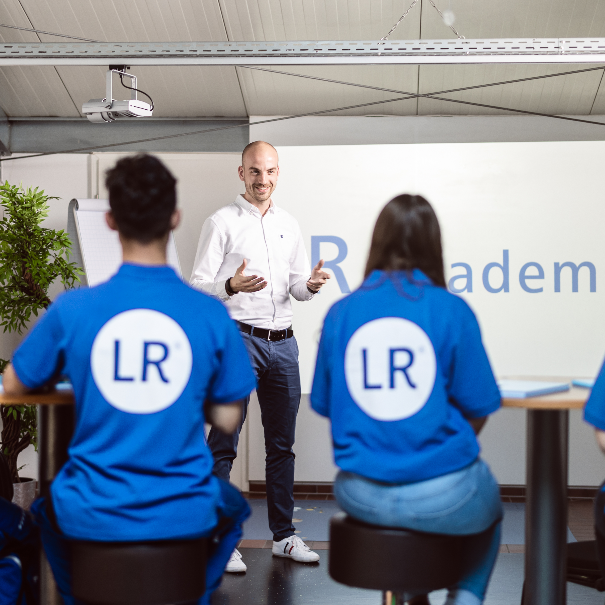 LR Akademie: Mitarbeiter steht vor großer Leinwand und präsentiert eine Folie auf der groß "LR Akademie" als blauer Schriftzug auf weißem Hintergrund sichtbar ist. Der Mitarbeiter steht gegenüber einer Gruppe von drei sitzenden Mitarbeitern, die alle mit dem Rücken zur Kamera in einer Reihe an Stehtischen auf Hockern sitzen und dem präsentierenden Mitarbeiter zuhören.