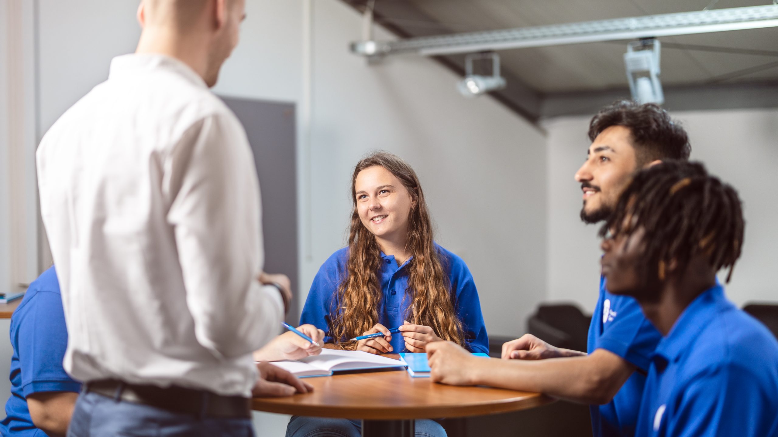 Workshop: Mitarbeiter in weißem Hemd unterhält sich am Tisch angeregt mit vier Mitarbeitern aus dem gewerblichen Bereich. Die Mitarbeiter machen sich Notizen.