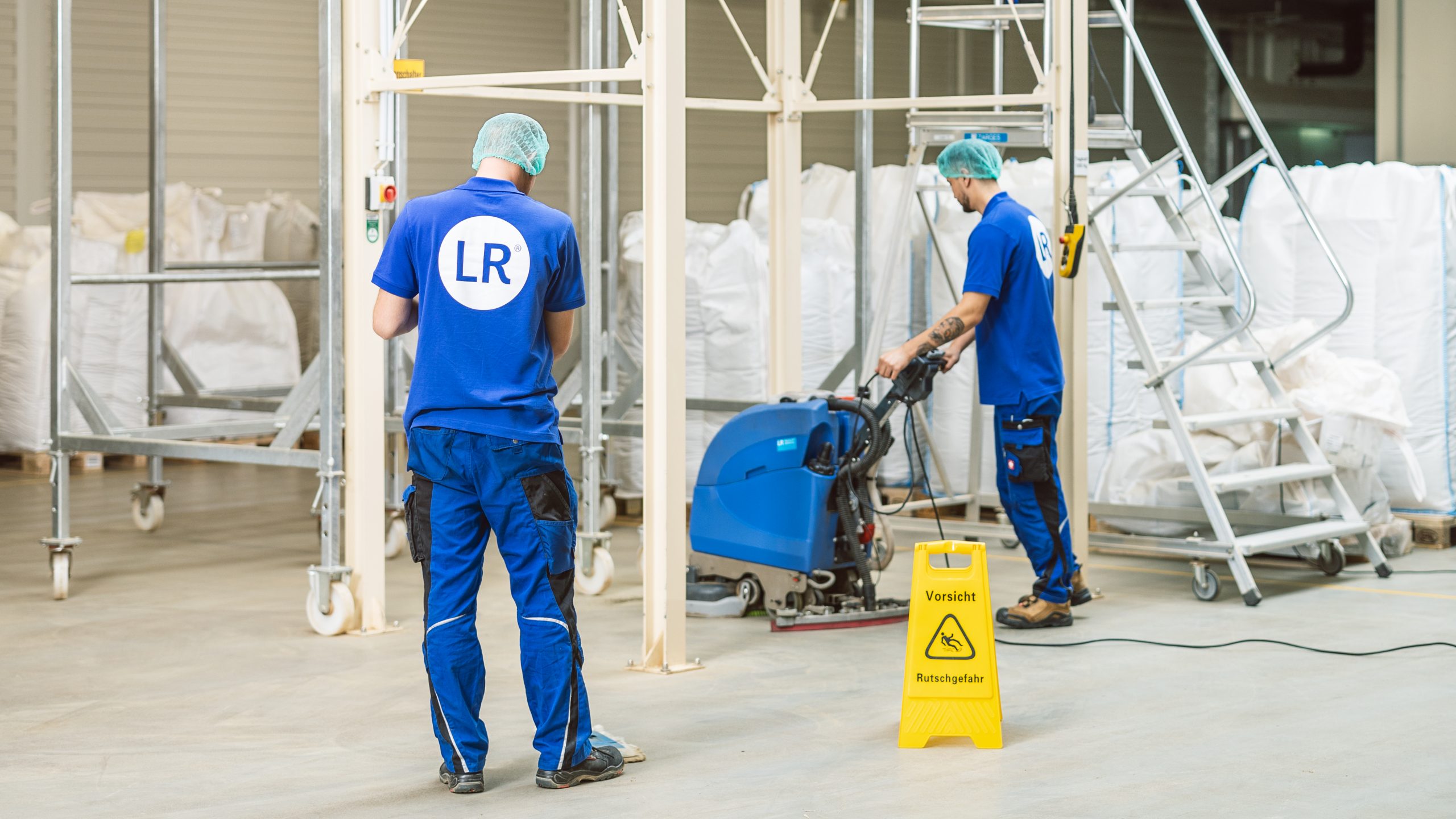 Zwei Mitarbeiter reinigen Fabrikhalle; alle tragen blaue Arbeitskleidung und Haarnetz; links: Mitarbeiter STEHT mit Rücken zur Kamera; rechts: Mitarbeiter reinigt Boden mit Maschine; In der Umgebung sind verpackjte Produkte des Unternehmens auf Holzpaletten zu sehen sowie ein Metallgerüst