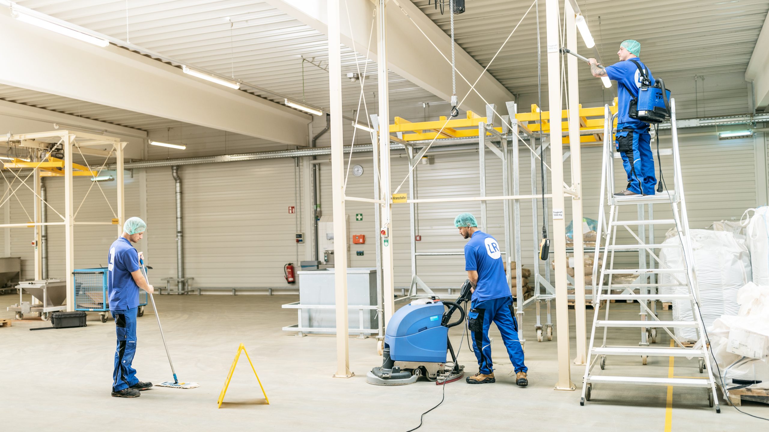 Drei Mitarbeiter reinigen Fabrikhalle; alle tragen blaue Arbeitskleidung und Haarnetz; links: Mitarbeiter wischt boden; Mitte: Mitarbeiter reinigt Boden mit Maschine; rechts auf Leiter: Mitarbeiter saugt Metallstangen ab; In der Umgebung sind verpackjte Produkte des Unternehmens auf Holzpaletten zu sehen sowie ein Metallgerüst
