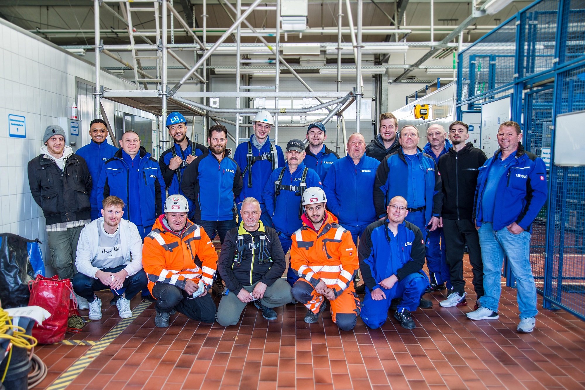 Gruppenfoto im LR Service Center: 19 Personen in unterschiedlicher Arbeitskleidung (teilweise mit Schutzhelmen) posiert in der Halle
